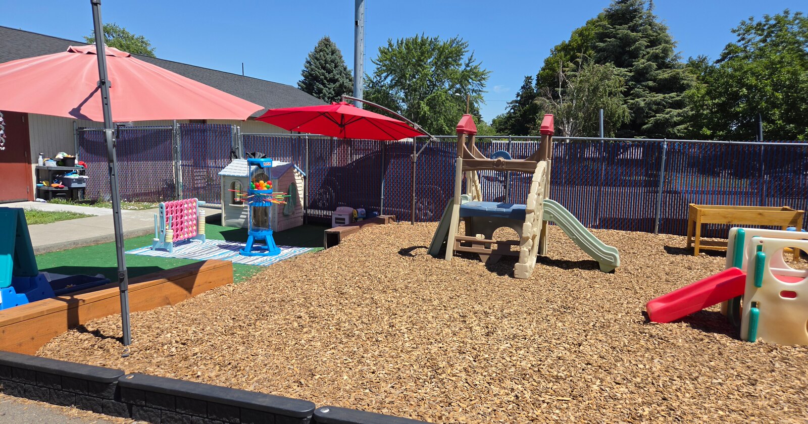 Apple Valley's fenced play yard with a climbing frame and slide, shaded seating, a playhouse and giant Connect Four